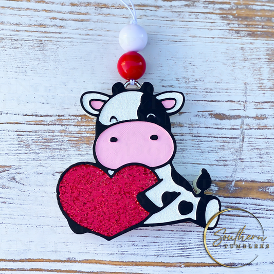 car air freshener in the shape of a cow holding a heart decorated with red and white glitter, pink and white paint. 